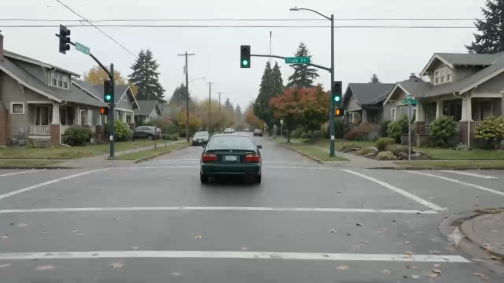 A rejected take seen from behind. A dark green sedan drives through the middle of an intersection on a two-way residential street, centered on the road rather than in its lane, under green traffic lights. It is a different car from the one in the film: its license plate sits high in the trunk lid between separate taillights, and the plate is not blurred. The street sign on the signal pole reads as garbled letters. Craftsman houses and autumn trees line both sides.