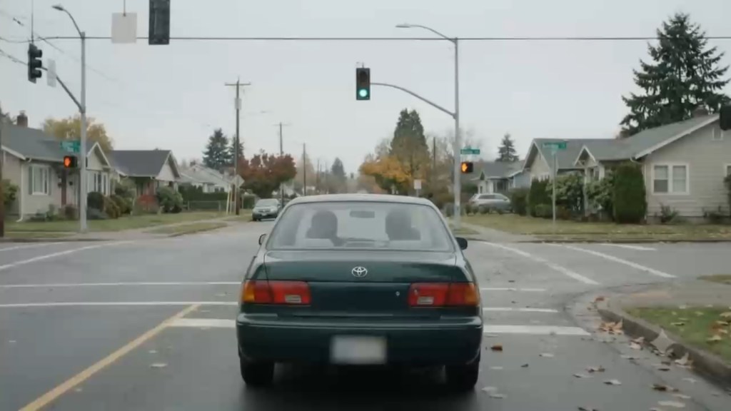 Seen from directly behind, the dark green sedan waits at an intersection under a green traffic light. The Toyota emblem is centered on the trunk and the license plate is blurred. Two figures sit in silhouette through the rear window, and houses and autumn trees line the street ahead.